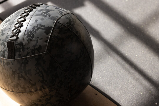 Camouflage medicine ball resting against plyo box on speckled rubber floor, catching sunlight