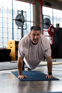 Middle-aged man holding push-up on blue mat in gym, wearing light shirt by barbell rack