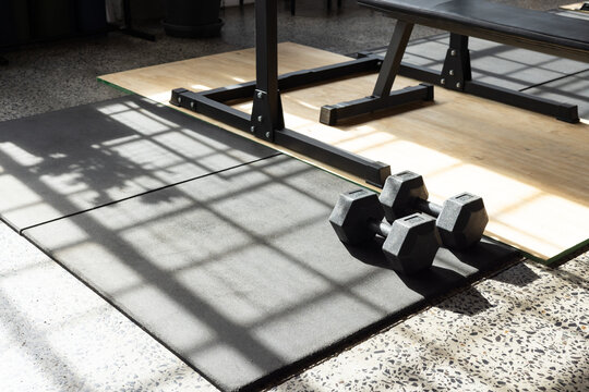 Pair of hex dumbbells are resting on black rubber mat near wooden platform, casting grid shadows