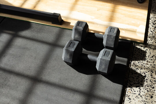 Hex dumbbells resting on black mat near window and wooden lifting platform, shadows crossing