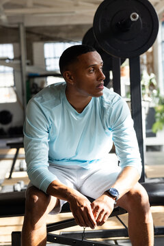 African American man sitting on flat bench at gym wearing blue top, barbell behind, watch visible