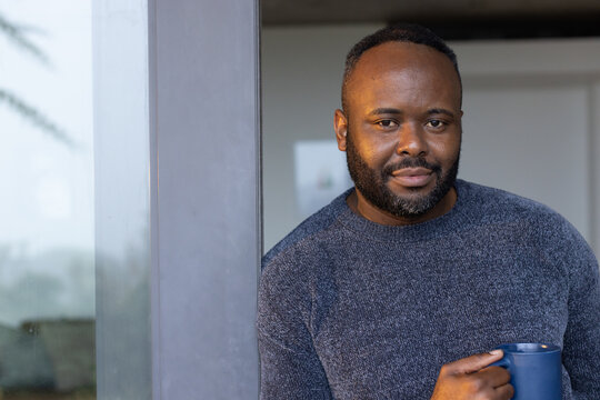 African man standing by glass sliding door, holding navy mug and wearing blue knit sweater