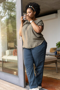 African American woman standing in sliding glass doorway holding ceramic mug and gazing left