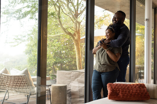 African couple standing at sliding glass doors, man embracing woman holding dark mug, copy space