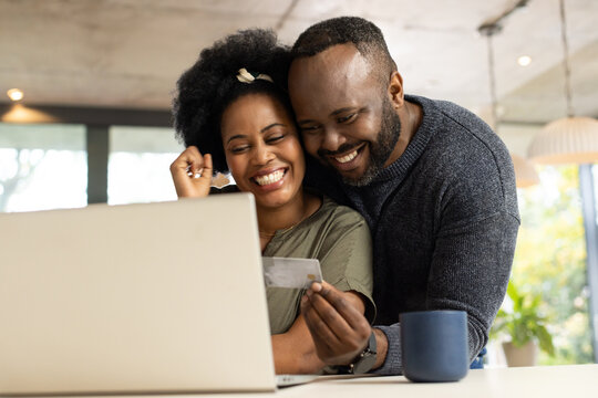 African American couple sitting at dining table using laptop, woman holding credit card, mug nearby