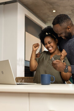 African American couple cheering, holding credit card and smartphone at kitchen counter with laptop