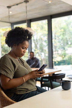 African woman sitting left cafe table using smartphone in olive blouse, ceramic mug, copy space