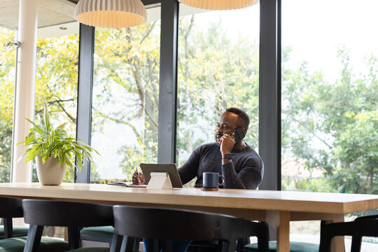 African man in dark sweater talking on smartphone and using tablet at communal table, copy space