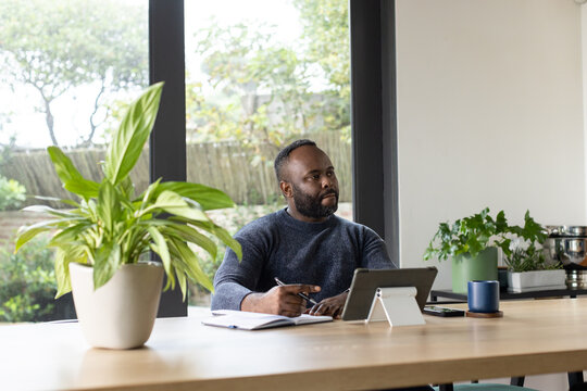 African man sitting at wooden table in home, writing in notebook and using tablet
