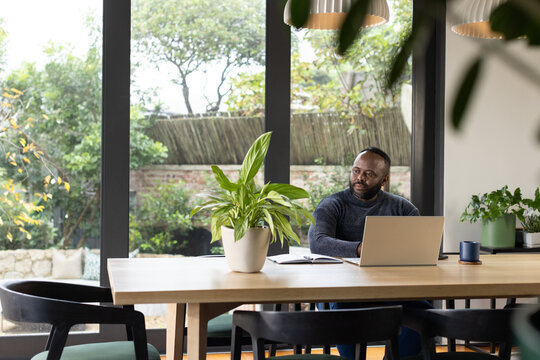 African American man working at wooden table in home using laptop, open notebook, mug, copy space