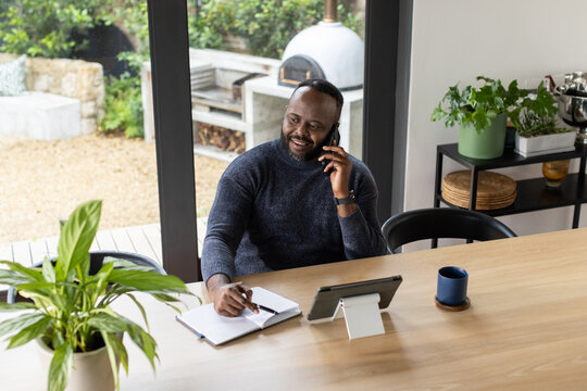 Adult African man sitting at home wearing gray sweater holding phone and tablet writing in notebook