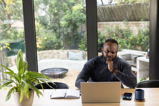 African American man working on laptop at wooden table by glass doors with notebook, mug, phone