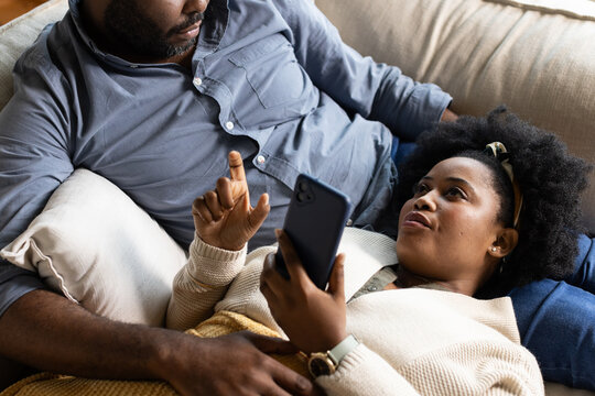 African American couple reclining on light sofa, woman holding smartphone and wearing cream sweater