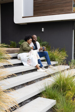 African couple sitting on front steps of modern house holding ceramic mugs, wearing sneakers