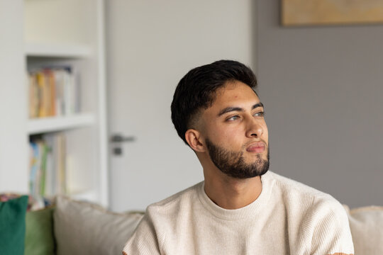 Adult male sitting on light-colored sofa wearing beige knit sweater, gazing right toward bookshelf
