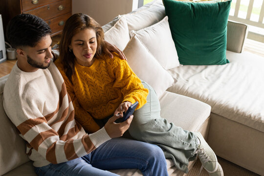 African American couple sitting on beige sofa in living room, holding smartphone, wearing sweaters