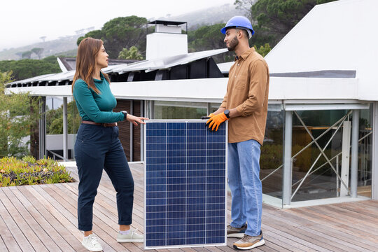 African American installer and client holding solar panel wearing blue helmet orange gloves on roof