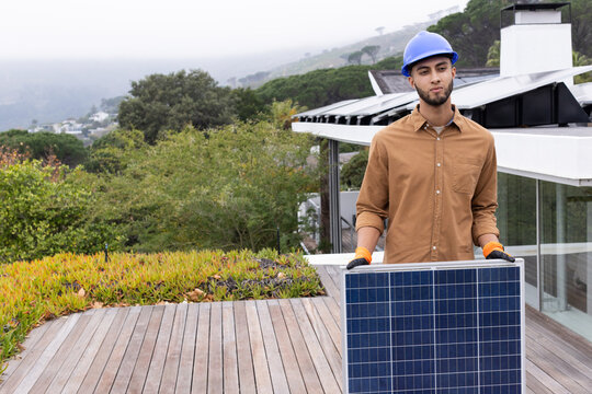 African American man holding solar panel on deck wearing blue hard hat, orange gloves, copy space