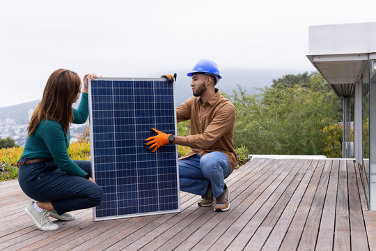 African American duo kneeling fitting solar panel on roof deck wearing blue hard hat orange gloves