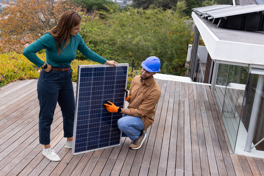 African American owner and tech holding solar panel on roof deck with blue helmet, orange gloves