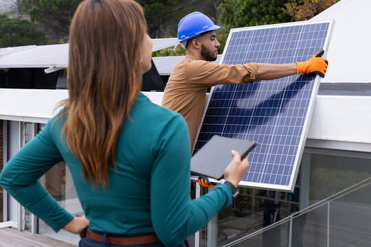 African American woman holding tablet and man lifting solar panel on roof wearing orange gloves
