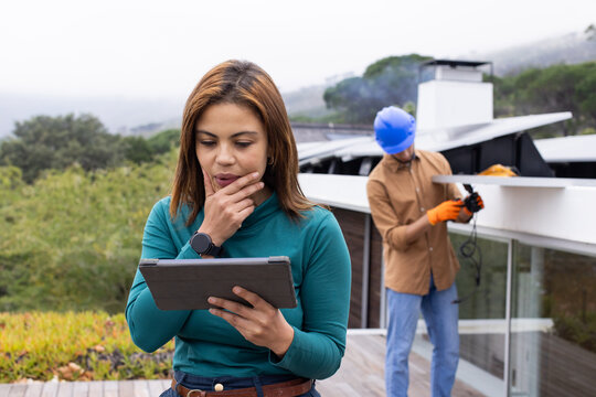 Woman manager holding tablet, reviewing solar on roof while male tech in hard hat handling cables