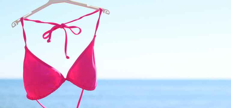 Bright pink bikini top hanging on a plastic hanger against a blurred blue sea and clear sky. Pink bikini close-up against the ocean on a sunny day. Concept of seaside resort, vacation