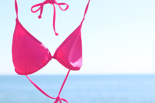 Bright pink bikini top hanging on a plastic hanger against a blurred blue sea and clear sky. Pink bikini close-up against the ocean on a sunny day. Concept of seaside resort, vacation