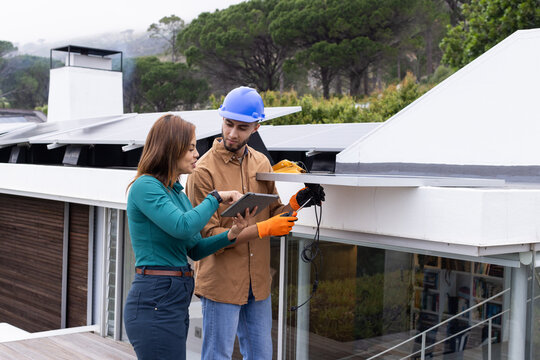African American team wearing hard hats installing solar panels on roof holding tablet and wiring