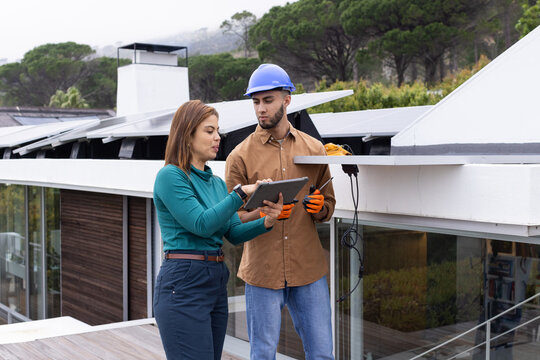 African American team wearing hardhat inspecting solar panels on roof with tablet and holding panel