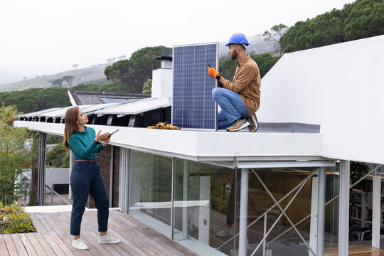 African American man and woman kneeling and standing on roof handling solar panel and tablet