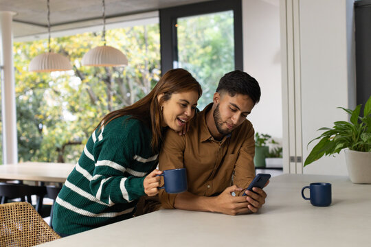 African American couple leaning on long light-colored countertop, sharing smartphone and blue mugs