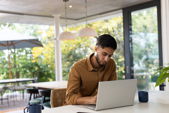 Man typing on silver laptop at dining table in modern home, blue mug under pendant lights