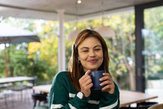 Adult African woman sitting in sunroom at table holding dark blue mug, wearing green sweater, watch