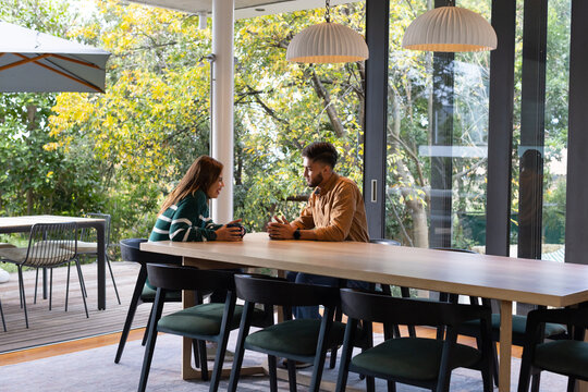 African American couple sitting at wooden dining table, holding cups, talking near sliding doors