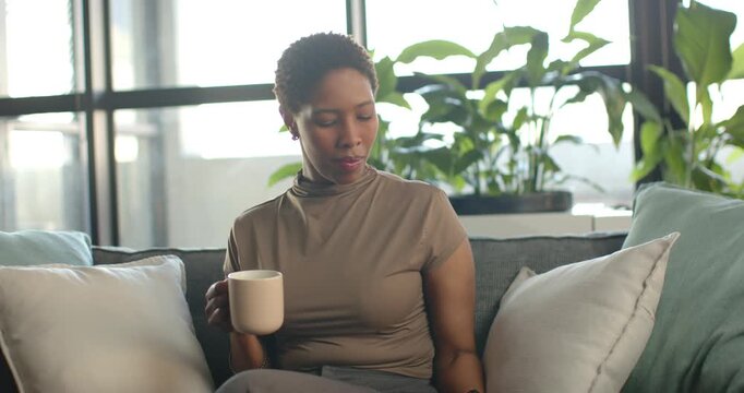 Woman glancing at smartphone, resting it, sipping mug on sofa near window, taking break