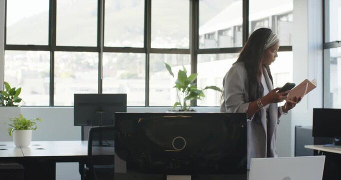 African American woman in coat reading phone, comparing with notebook, moving to window, copy space