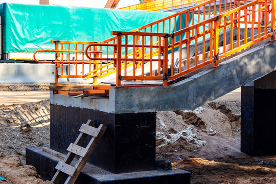 Bright orange metal railings outline a concrete pedestrian bridge under construction above a highway, featuring modern urban infrastructure elements and raw building site materials