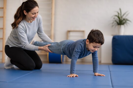 Woman therapist helping young boy do wheelbarrow exercise on mat during sensory integration therapy. Physical activity supporting attention and focus for child with ADHD and autism spectrum disorder