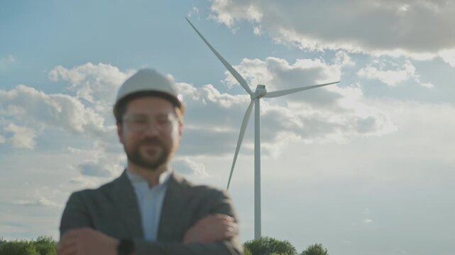 Man in hard hat and glasses with arms crossed smiles in front of wind turbine on sunny day. Close-up portrait of successful engineer promoting sustainable technology
