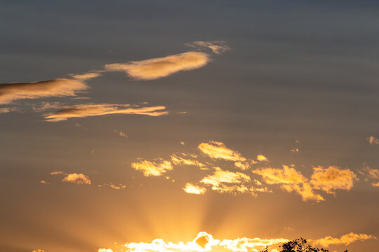 Golden hour sky featuring radiant sunbeams breaking through soft, glowing clouds, creating a peaceful and serene atmosphere during a vibrant late afternoon sunset over a silhouette horizon