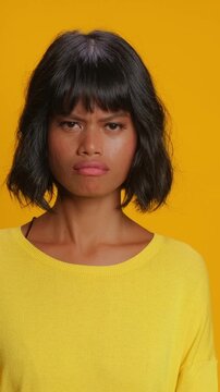Upset Southeast Asian woman looks at camera with disappointed face and shakes head. Emotional person shows negative reaction and sadness while standing against bright yellow studio background.