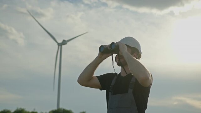 Man in hard hat and overalls uses binoculars to observe wind turbine against cloudy sky, representing renewable energy sector. Engineer monitoring sustainable technology infrastructure
