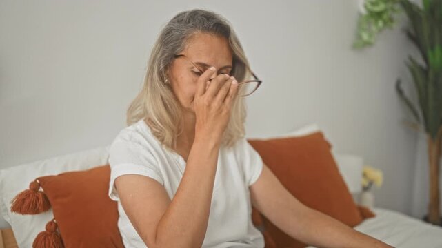Woman pinches bridge of nose while seated on bed in a bedroom with rust pillows and white sheets; fatigue reflection.