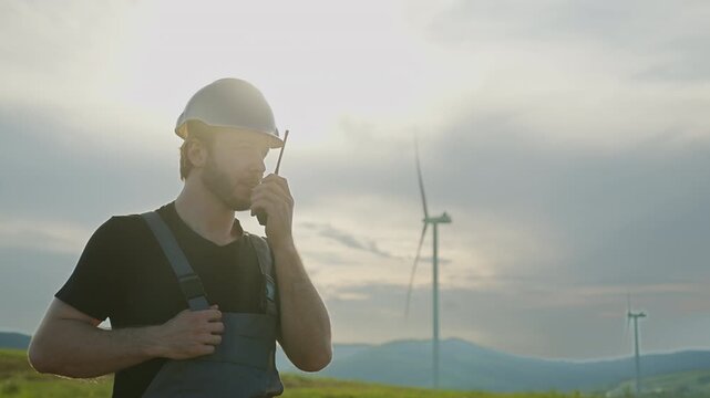 Man in hard hat and overalls speaks into radio with wind turbines in background during daytime. Footage illustrates field communication in renewable energy site