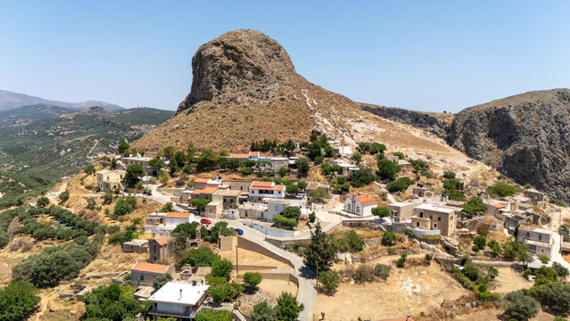Drone view of a small village at the base of a steep rocky hill on Crete, showing traditional stone houses, winding roads, and sparse vegetation under clear blue sky.