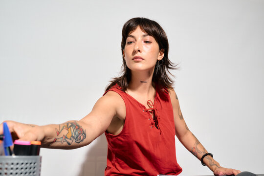 A woman with tattoos and a red top reaches toward a pen holder. Picking up a pen while working in the office