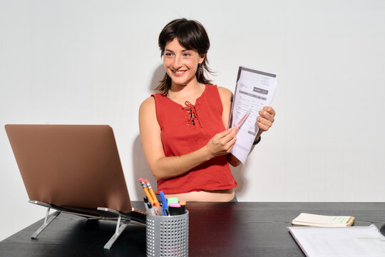 Woman Showing a CV during a job video call. She smiles confidently while holding papers near a laptop. 
