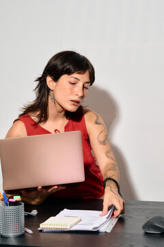 A focused woman works at her desk with laptop and papers. She&rsquo;s deeply concentrated. Picking up business papers while holding her laptop at office