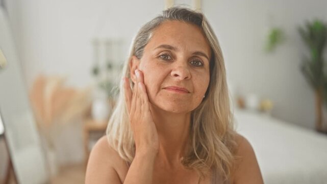 Woman touching cheek with hand while seated on bed in a bright bedroom, closeup skincare gesture and reflective gaze; serenity.